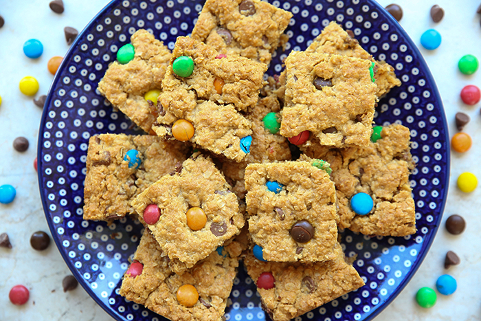 A white polka dotted dark blue plate of square-cut monster cookie bars with m&ms and chocolate chips surrounding the plate.