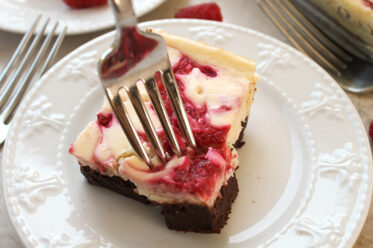 A raspberry cheesecake brownie on a small white plate with a metal fork inserted downward into a corner of the brownie. In the upper left is a fork head a part of another small plate.