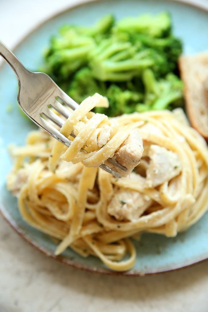 A plate with cooked broccoli, sliced bread, and a scoop of lemon chicken pasta with a fork held above wrapped with noodles and chicken.