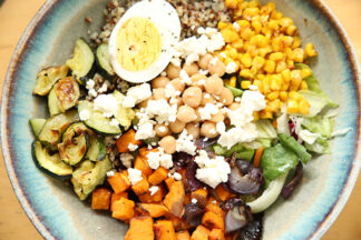 A bowl filled with quinoa, roasted corn, zucchini, chopped sweet potato and spinach topped with feta cheese, half an hard-boiled egg, chickpeas, and balsamic dressing.