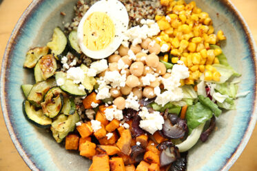 A bowl filled with quinoa, roasted corn, zucchini, chopped sweet potato and spinach topped with feta cheese, half an hard-boiled egg, chickpeas, and balsamic dressing.