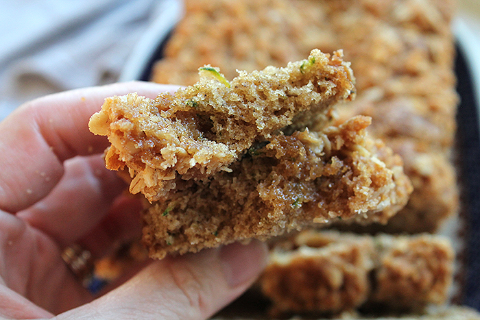 A slice of zucchini bread with streusel broken in half held above a sliced loaf of zucchini bread on an oval platter.
