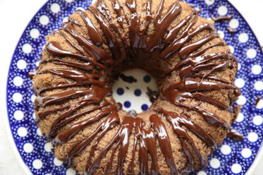 A whole chocolate zucchini spice bundt cake with chocolate ganache drizzled down the sides and in the middle sitting on a round Polish pottery plate with white polka dots and a dark blue background.