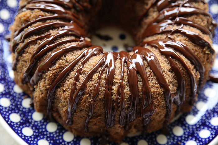 A closeup picture of a chocolate zucchini bundt cake drizzled with chocolate ganache sitting on a round dark blue Polish pottery plate with white polka dots.