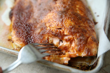 The corner of a baking sheet with parchment paper with a filet of sweet and smoky brown sugar salmon and a fork inserted into the salmon from the left.