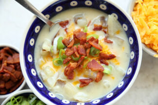 A dark blue white polka dotted soup bowl of loaded baked potato soup topped with sliced green onions, shredded cheddar cheese, and bacon crumbles. In the upper right corner is a partial picture of a bowl of shredded cheese, and in the bottom left corner is a partial picture of a bowl of bacon pieces and a bowl of sliced green onions.