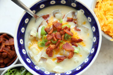 A dark blue white polka dotted soup bowl of loaded baked potato soup topped with sliced green onions, shredded cheddar cheese, and bacon crumbles. In the upper right corner is a partial picture of a bowl of shredded cheese, and in the bottom left corner is a partial picture of a bowl of bacon pieces and a bowl of sliced green onions.