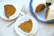 Two slices of pumpkin bundt cake with cream cheese frosting sitting on small plates with two forks crisscross on each other between the plates. In the upper right corner is a pumpkin bundt cake with cream cheese frosting on a round platter with a couple slices cut out.