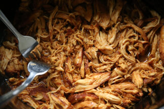 A crockpot of shredded honey garlic chicken with two forks inserted into the chicken on the left.