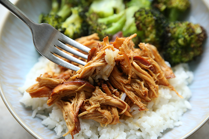 Shredded garlic honey chicken served over rice with a fork inserted diagonally on the left and a serving of steamed broccoli all on a plate.