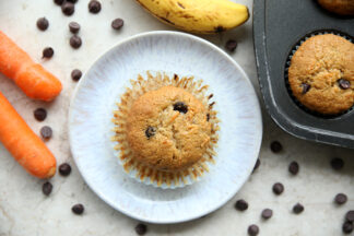 A banana carrot chocolate chip muffin with the wrapper pealed down sitting on a small plate with chocolate chips scattered around the plate. On the left of the plate are two diagonally placed carrots. Above the plate is a partial banana and to the right is a muffin sitting in a muffin pan.