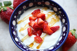 A dark blue Polish pottery bowl with white polka dots with homemade yogurt drizzled with honey and topped with slice strawberries. Two strawberries are in the upper left corner and one in the bottom right corner.
