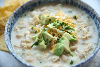 White chicken chili with hominy in soup bowl and topped with shredded Colby Jack cheese, chopped avocado and chopped cilantro with tortilla chips on the upper left.