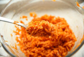 A glass mixing bowl with carrot kimchi with a spoon held above with a scoop of carrot kimchi on it.