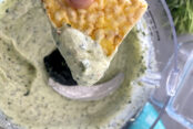 A food processor with cilantro ranch protein dip in it and triangular corn chip with dip on the tip being held above the food processor.