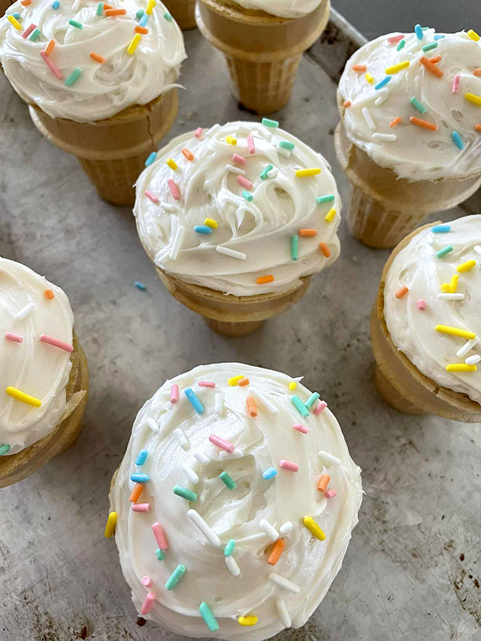 Vanilla frosted cupcake cones with sprinkles sitting on a large baking sheet.