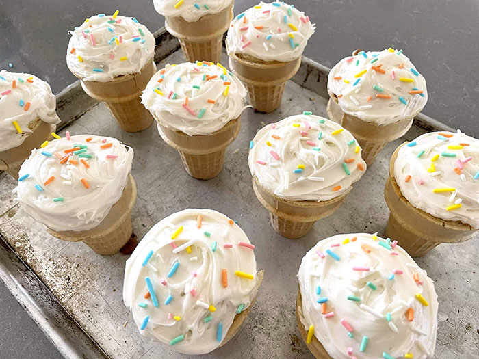 A large baking sheet with cupcake cones frosted with vanilla frosting and topped with multicolored sprinkles.