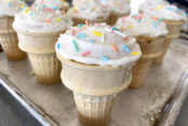 Ice cream cone cupcakes with vanilla frosting and sprinkles on a cookie sheet.