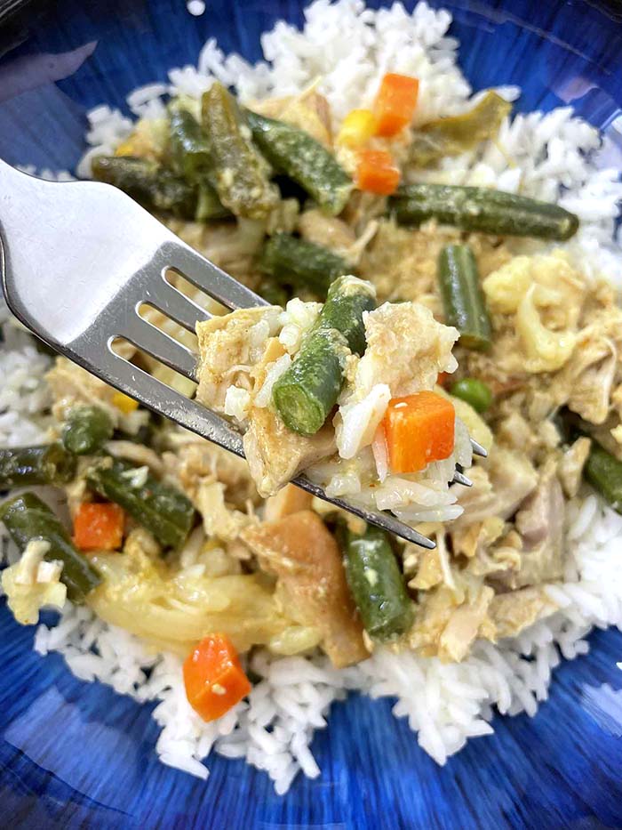A dark blue dinner plate with white rice topped with chicken curry mixed with green beans, carrots, peas and corn.