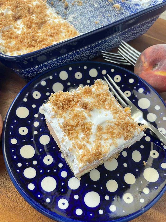 A square slice of no-bake fresh peach dessert on a small Polish pottery plate with a dark blue background and white polka dots. A fork is lying next to the slice. Above is a partial picture of a dark blue patterned 9x13 Polish pottery dish of fresh peach dessert with a piece cut out of the corner. On the upper right is a whole peach and two fork heads.