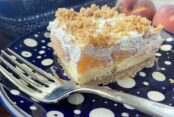A slice of fresh peach dessert sitting on a small dark blue plate with polka dots with a fork on the plate. In the background is the edge of patterned dark blue 9x13 dish and two whole peaches.