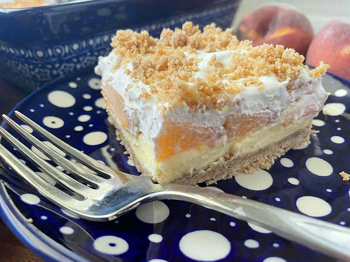A slice of fresh peach dessert sitting on a small dark blue plate with polka dots with a fork on the plate. In the background is the edge of patterned dark blue 9x13 dish and two whole peaches.