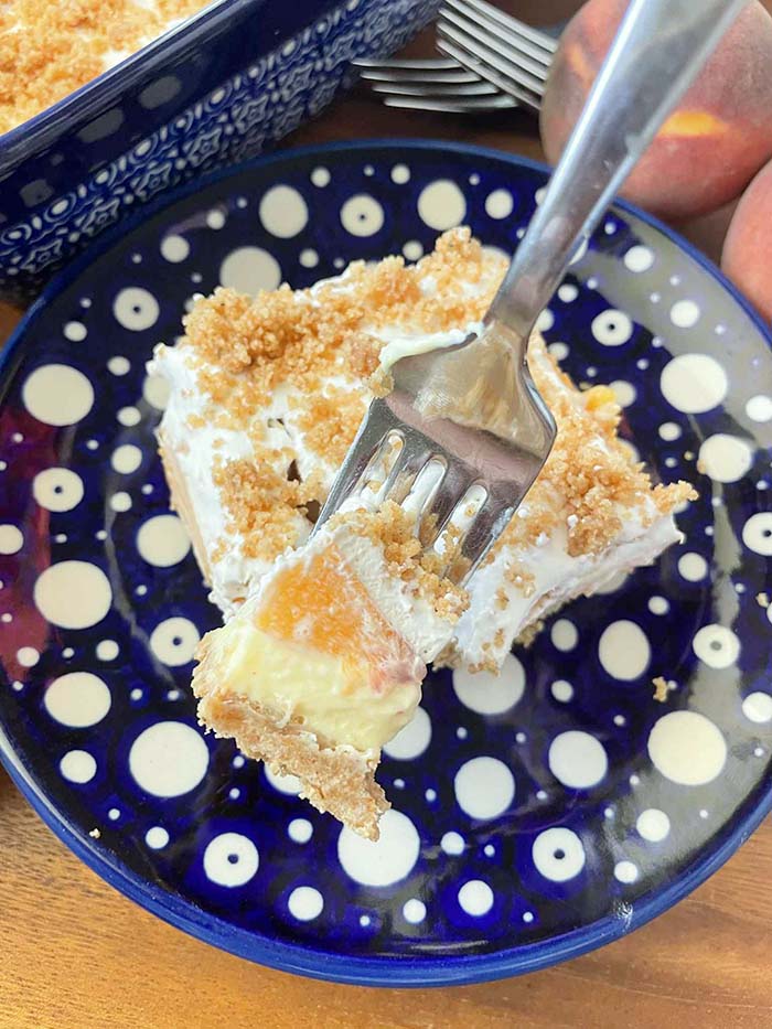 A small dark blue plate with white polka dots with a slice of fresh peach no-bake dessert and a fork held diagonally inserted into the corner of the dessert lifting a bite up. In the background are two peaches, two fork heads, and the edge of a 9x13 baking dish.