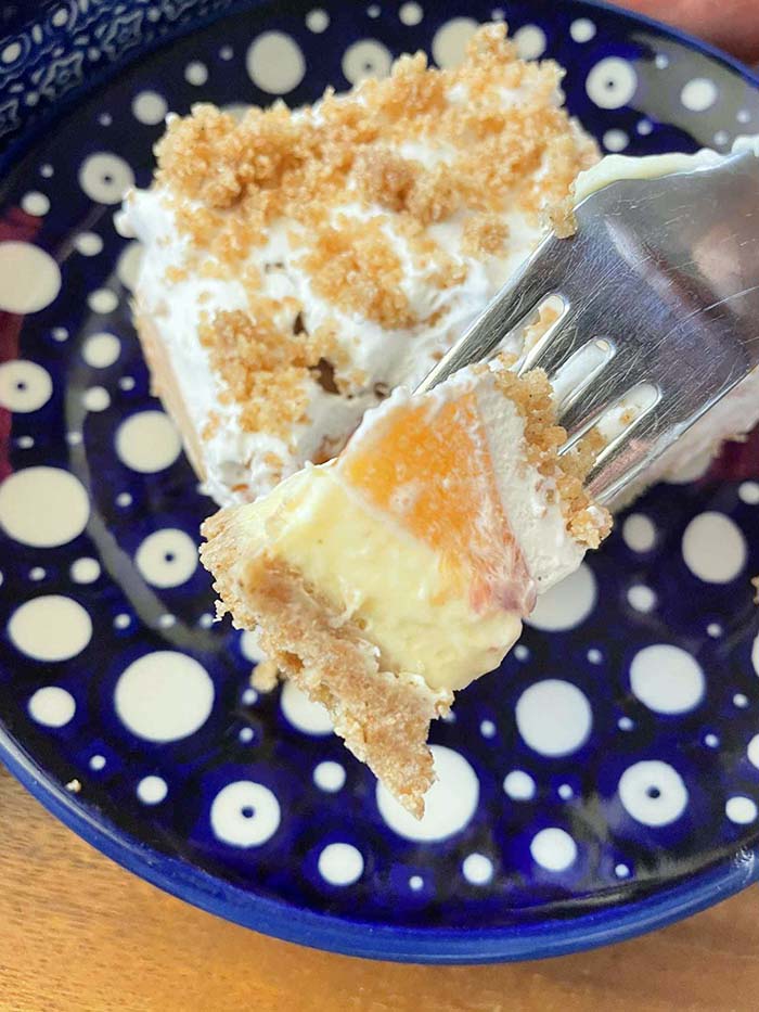 A fork head with a piece of fresh peach dessert held above a white polka dotted dark blue partnered plate with a square slice of fresh peach dessert.