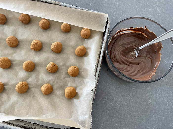 A parchment-lined baking sheet of peanut butter balls with a small glass bowl of melted semi-sweet chocolate with a spoon in it on the right.