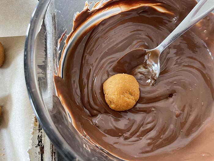 A glass bowl of melted semi-sweet chocolate with a spoon inserted from the right and a peanut butter ball on top of the chocolate next to the spoon.