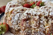 A strawberry rhubarb bundt cake dusted with powdered sugar with whole strawberries in the middle sitting on a round plate with a couple of whole strawberries on the left.