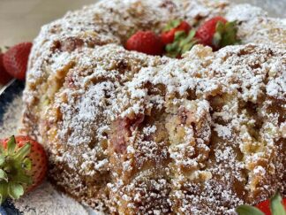 A strawberry rhubarb bundt cake dusted with powdered sugar with whole strawberries in the middle sitting on a round plate with a couple of whole strawberries on the left.