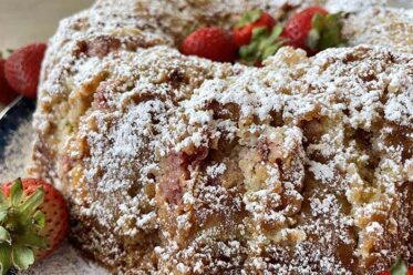 A strawberry rhubarb bundt cake dusted with powdered sugar with whole strawberries in the middle sitting on a round plate with a couple of whole strawberries on the left.