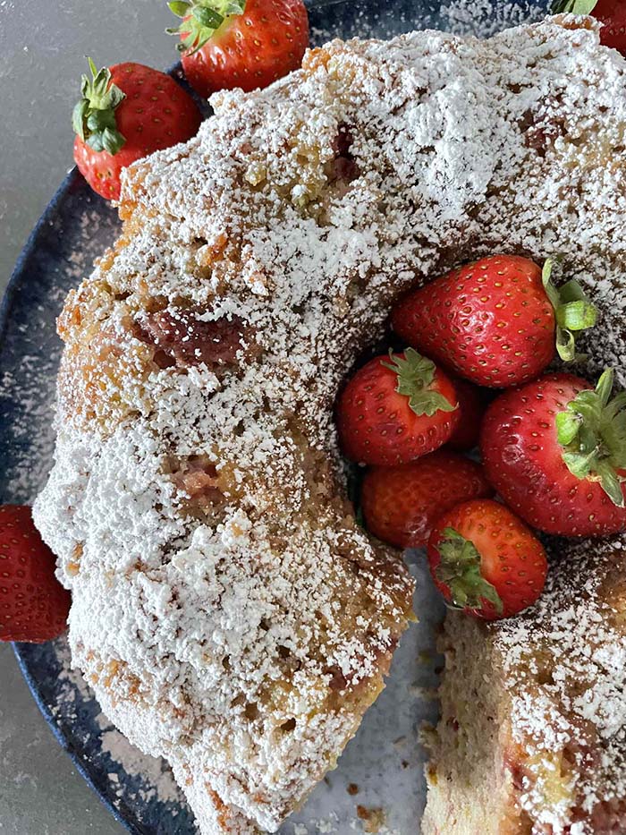 Strawberry Rhubarb Bundt Cake dusted with powdered sugar with a slice cut out sitting on a large round plate with some whole strawberries on the edge. Several whole strawberries are in the hole in the middle of the cake.