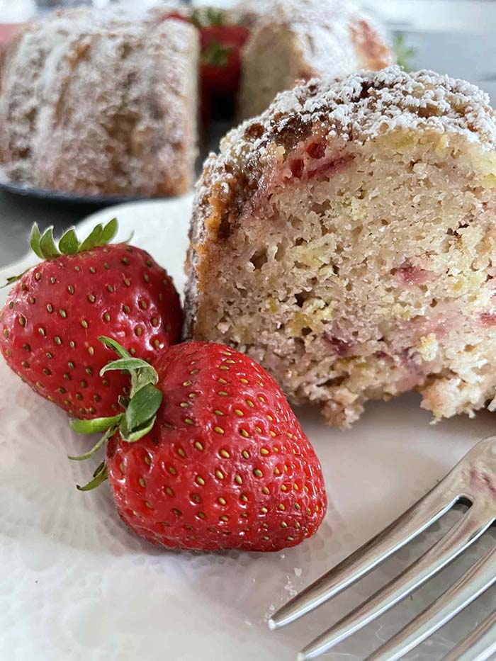 A slice of strawberry rhubarb bundt cake on a small plate with two whole strawberries and a fork head. In the background is a strawberry bundt cake with a slice cut out of it.