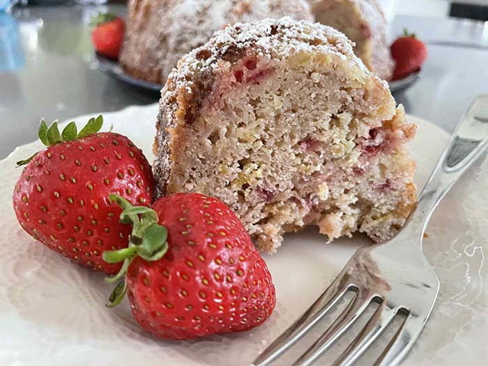 Two whole strawberries sitting next to a slice of strawberry rhubarb bundt cake and a fork on a small white plate.