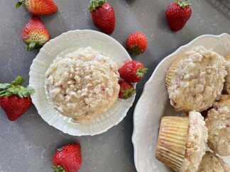 A strawberry rhubarb muffin on a small plate with two whole strawberries on the left. A partial picture of a plate bowl with strawberry rhubarb muffins. Whole strawberries are scattered around the small plate.