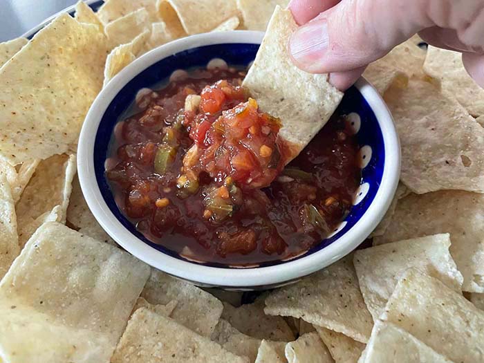 Tortilla chips surrounding a bowl of homemade salsa with a hand holding a tortilla chip with salsa on it above the bowl of salsa.