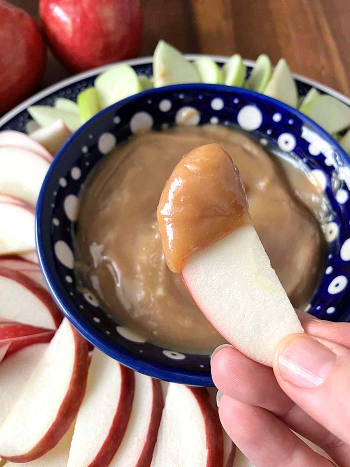 An apple slice with caramel dip on the tip held above a round plate with green and red apple slices with a dark blue and white polka-dotted small bowl in the middle with caramel sauce.