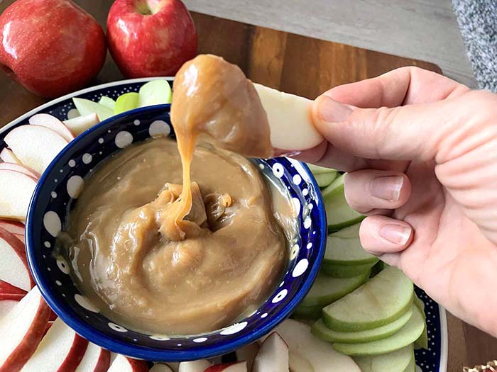An apple slice with caramel sauce on the tip and dripping into a small dark blue and white polka-dotted bowl of caramel on a large round plate with sliced green and red apples surrounding the small bowl.