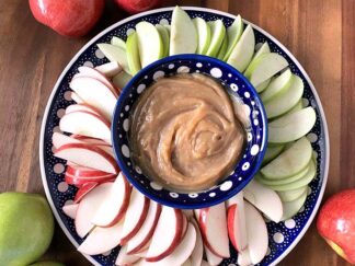 A large round plate with sliced red and green apples with a dark blue and white polka-dotted patterned small bowl in the middle with caramel dip. Surrounding the plate are apples in the upper left corner, bottom left corner, and bottom right corner.