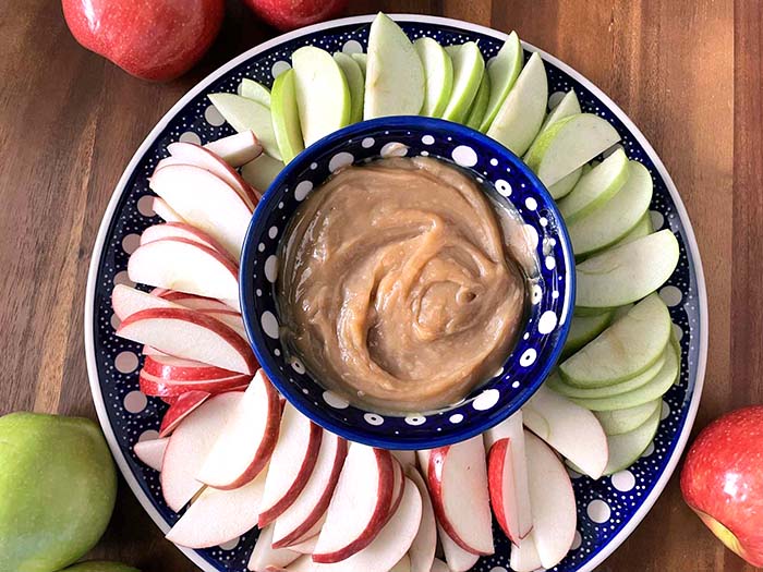 A large round plate with sliced red and green apples with a dark blue and white polka-dotted patterned small bowl in the middle with caramel dip. Surrounding the plate are apples in the upper left corner, bottom left corner, and bottom right corner.