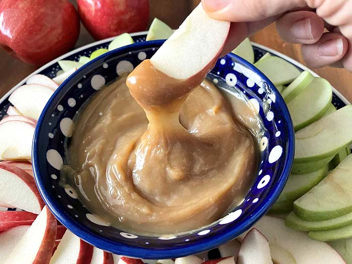 A large round plate with red and green sliced apples with a small dark blue and white polka-dotted bowl of caramel apple dip. A red apple slice is held above the small bowl with caramel lifting out and on the tip of the apple.