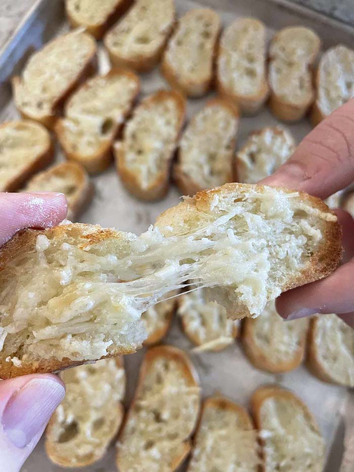 A slice of toasted parmesan baguette being torn apart held above a sheet pan full of toasted parmesan baguette slices.