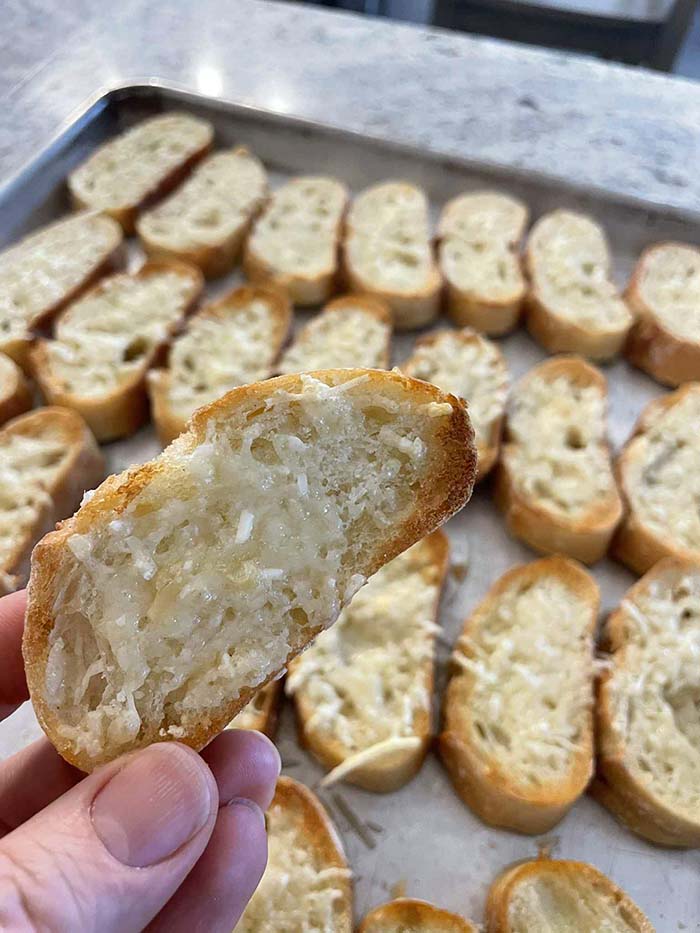 A toasted parmesan baguette slice held above a baking sheet full of sliced toasted parmesan baguette pieces.