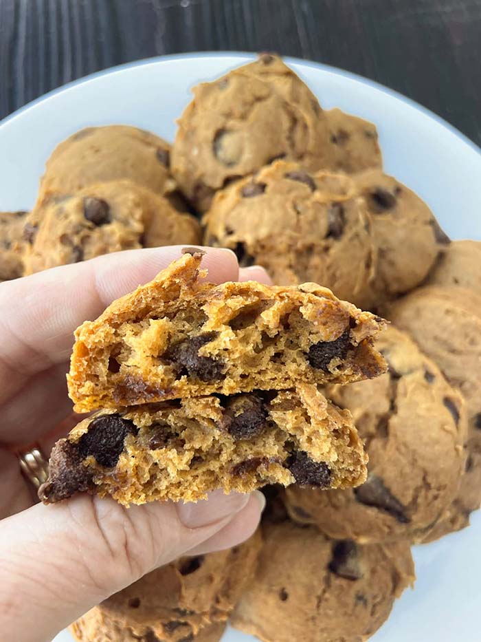 Two halves of a pumpkin chocolate chip cookie held above a plate of pumpkin chocolate chip cookies.