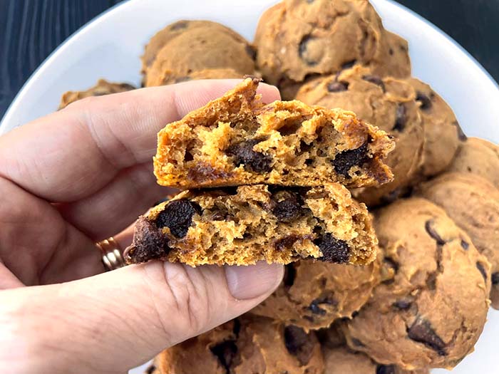 A plate of pumpkin chocolate chip cookies with a hand held above holding two cookie halves.