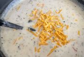 A large pot of Cheeseburger Soup with a sprinkling of shredded cheddar cheese on top in the middle with a black ladle inserted from the left.