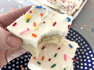 A square slice of sugar cookie cake frosted with sprinkles on top with a bite out of one corner held above a dark blue plate with white polka-dots with another slice of sugar cookie cake on it.