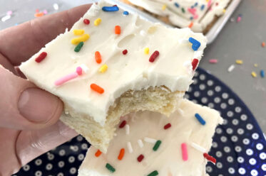 A square slice of sugar cookie cake frosted with sprinkles on top with a bite out of one corner held above a dark blue plate with white polka-dots with another slice of sugar cookie cake on it.